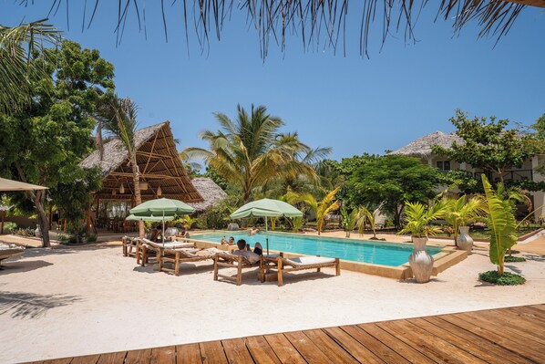A poolside area with lounge chairs and umbrellas, surrounded by palm trees and a thatched-roof building. - Hostel 197 (Nungwi)