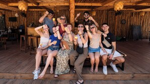 Un groupe de personnes est assis sur des bancs en bois, à l’abri sous un toit de chaume. L’intérieur est rustique, avec des éléments en bois apparents, et un panneau affichant le menu est visible.