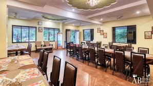 A spacious dining area with multiple tables and chairs, large windows with views of greenery, and ceiling fans. - Tiger King Resort (Chimur)