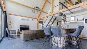 A spacious living area with a high ceiling, a central seating area with modern chairs, a television, and a kitchen with wooden cabinets and a staircase. - Thatched bungalow with private terrace (Baarle-nassau)