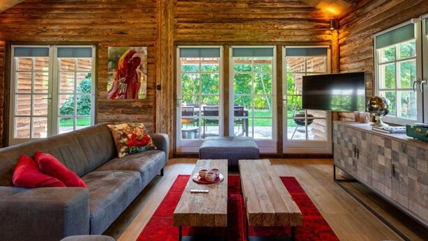 Interior de una cabaña de troncos con sofá, dos mesas de madera, televisor y un ventanal con vistas a la vegetación.