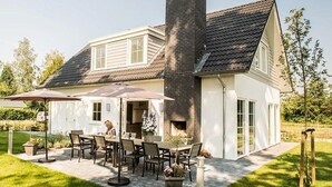 Une maison à deux étages dotée d’un patio comprenant une table et des chaises, une cheminée en briques et un jardin.