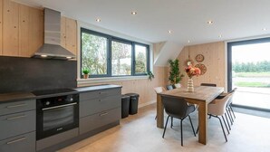 A modern kitchen with built-in appliances, a dining area with a wooden table and chairs, and large windows offering a view of the outdoors. - Seaside Holiday Home with Garden (Groote Keeten)