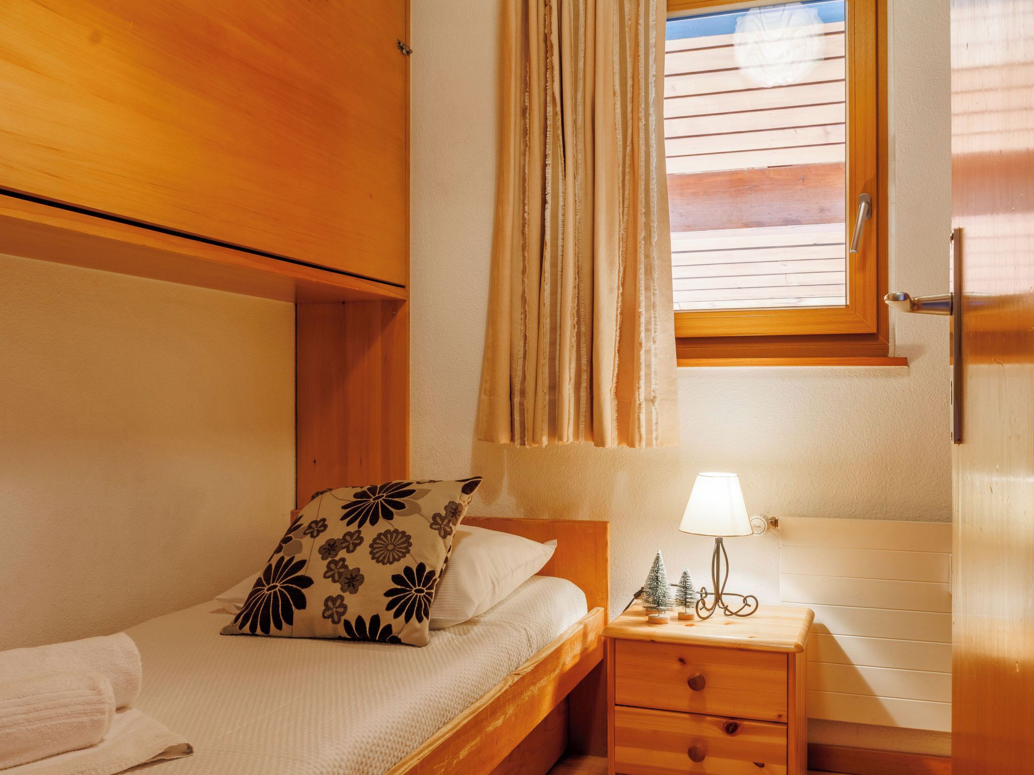 A bedroom with a wooden bed, a nightstand with a lamp, a window with curtains, and a wooden wardrobe.