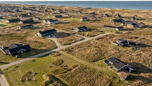 Vista aérea de un pueblo costero con casas, caminos y aerogeneradores a lo lejos.