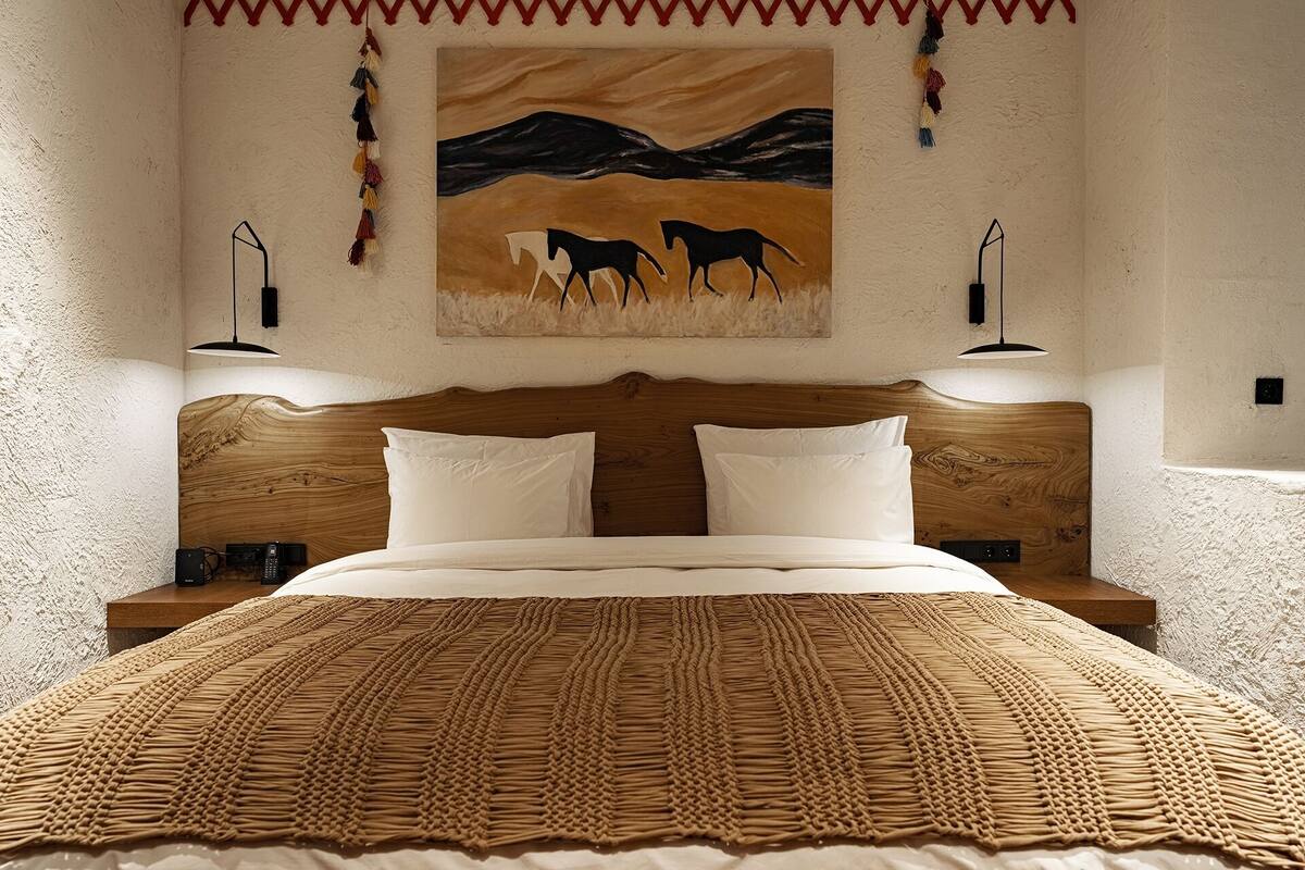 Une chambre à coucher avec une tête de lit en bois, deux tables de chevet, un grand lit recouvert d’une couverture à motifs, et un tableau représentant des chevaux accroché au mur.