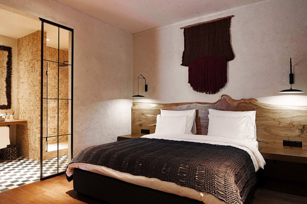 Une chambre à coucher avec un lit, une tête de lit en bois, une table de chevet, une lampe fixée au mur et un sol à carreaux.