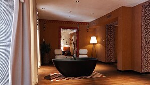 Una habitación con una bañera negra grande, suelo de madera, una alfombra estampada y una lámpara.