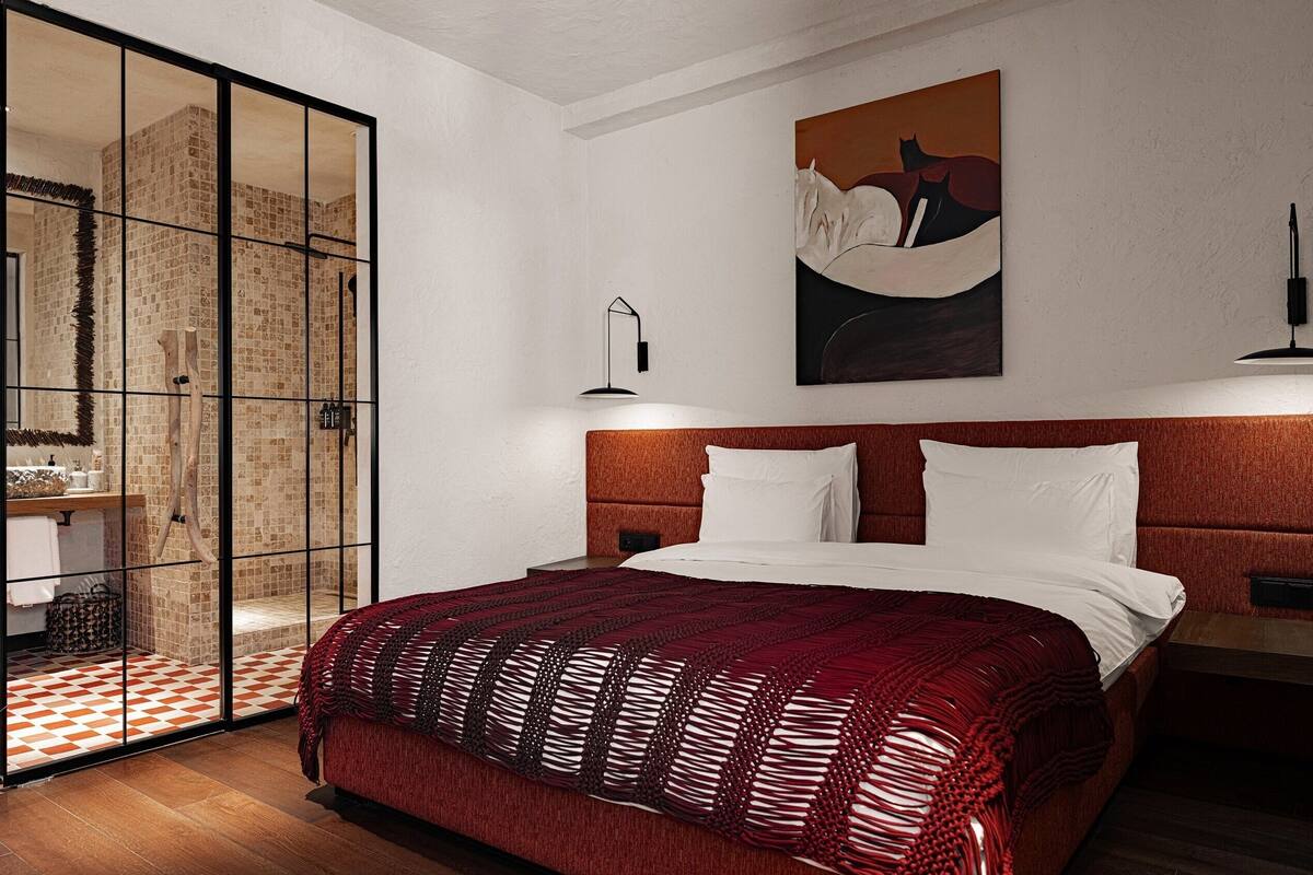Une chambre d’hôtel avec un grand lit, une tête de lit en bois, une couvre-lit à motifs rouges et blancs, un tableau au mur et une cabine de douche en verre.