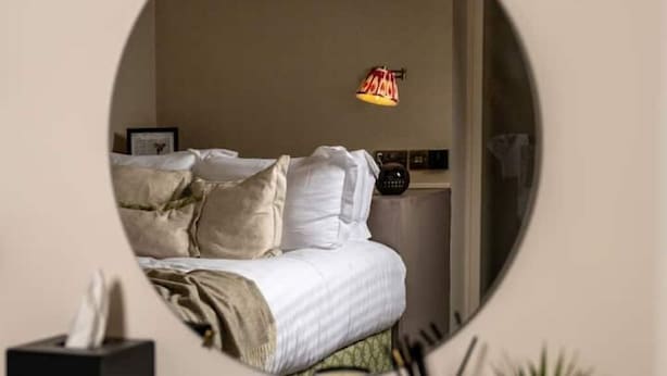 A bedroom with a bed, bedside table, lamp, and a mirror reflecting the room.