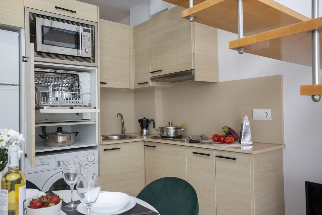A compact kitchen with a built-in dishwasher, microwave, and stove, a small dining area with a table and chairs, and a wooden ceiling structure.