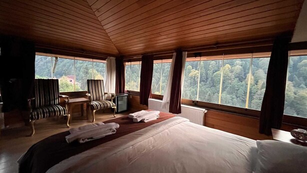 A wooden-paneled room with a bed, a desk, a chair, and a view of the forest through large windows. - Ayder Hotel  (Çamlıhemşin)