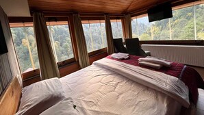 A hotel room with a wooden ceiling, a bed with white linens, two chairs, a TV mounted on the wall, and a view of a forest through large windows. - Ayder Hotel  (Çamlıhemşin)