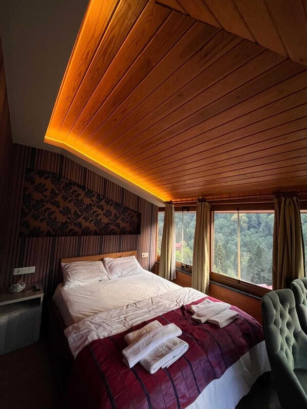 A bedroom with a wooden ceiling, a large bed with white bedding, a red and white quilt, and a view of the outdoors through a window. - Ayder Hotel (Çamlıhemşin)