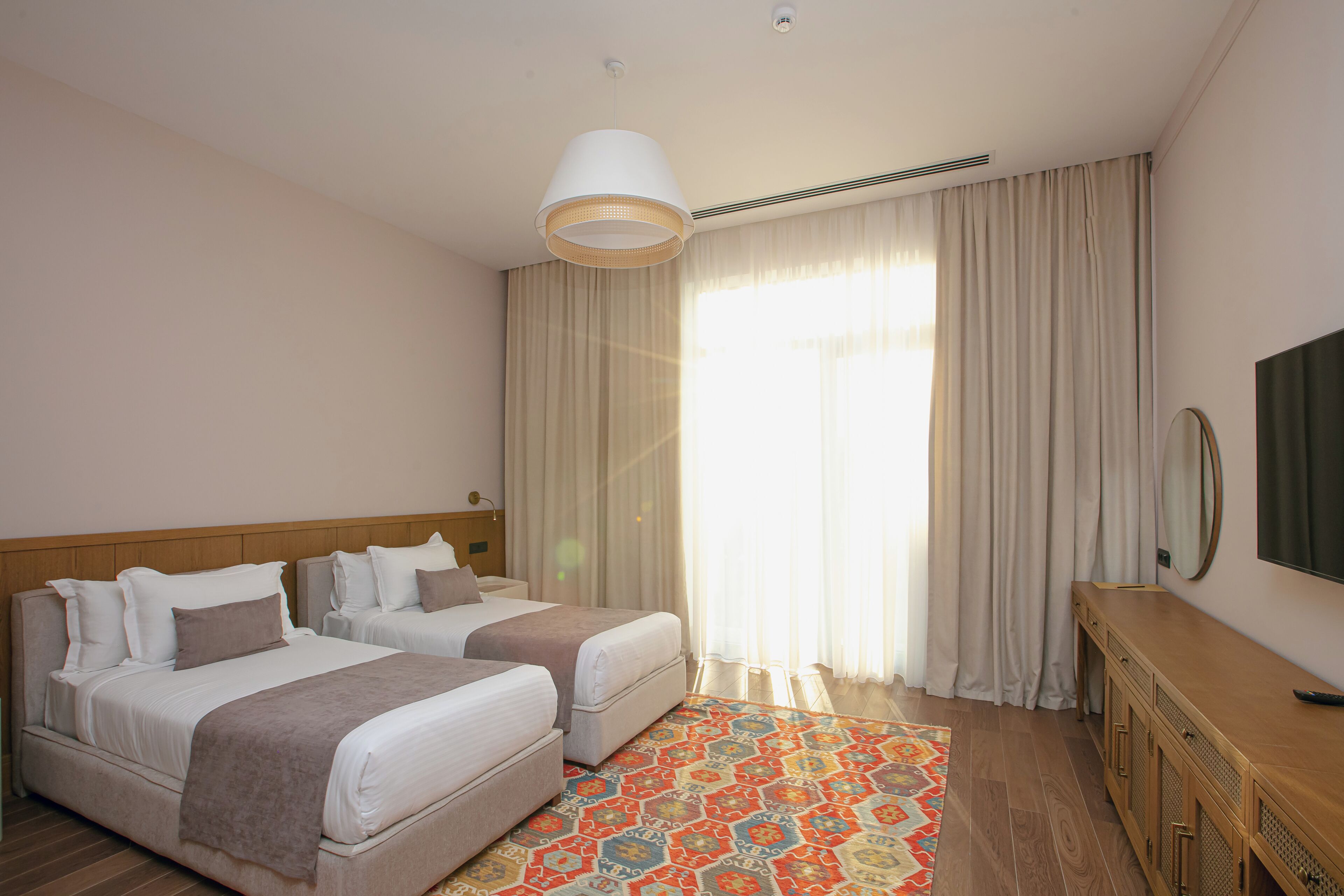 A hotel room with two beds, a wooden headboard, a large window with curtains, a television, and a patterned rug.