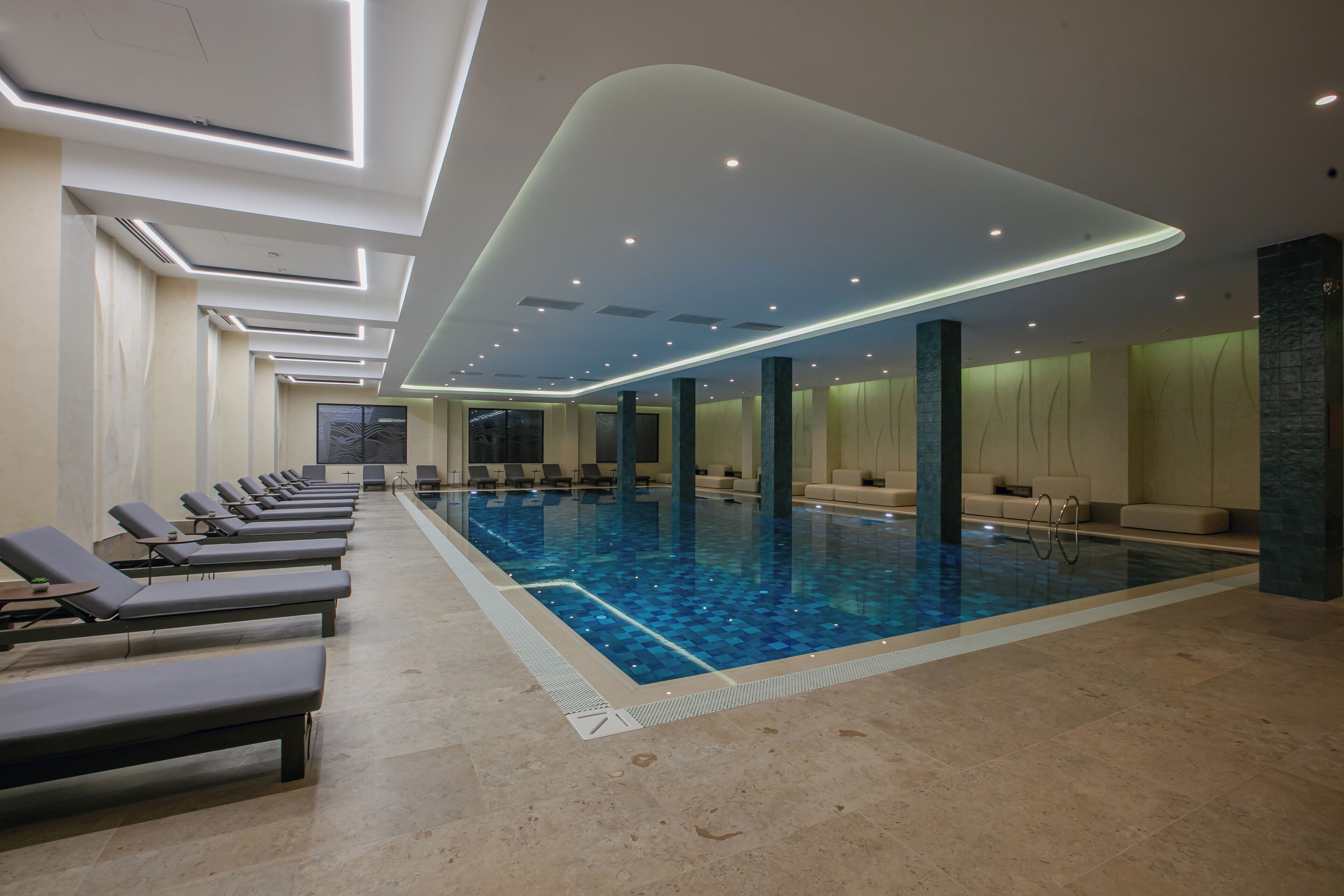 A modern indoor swimming pool with lounge chairs and a high ceiling with recessed lighting.