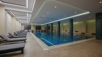 A modern indoor swimming pool with lounge chairs and a high ceiling with recessed lighting.