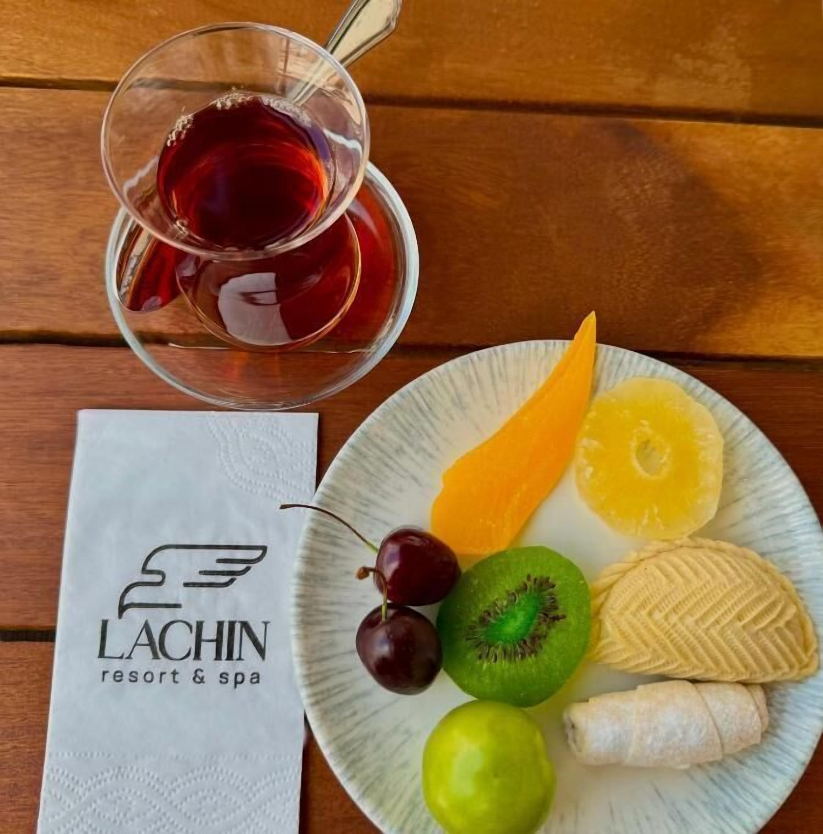 A plate with fruits and a glass of red beverage on a wooden table, next to a napkin with a logo.
