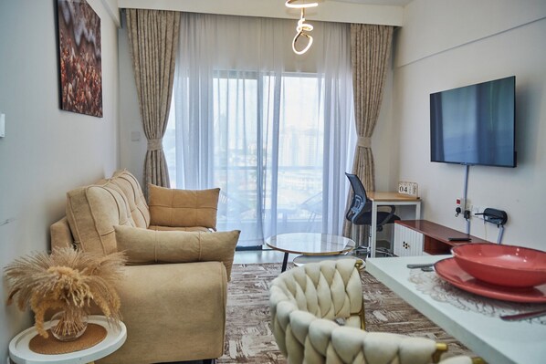 Ein modernes Wohnzimmer mit einer Couch, einem Esstisch mit einer roten Schüssel, einem Fernseher und Blick auf eine Stadtlandschaft durch große Fenster.