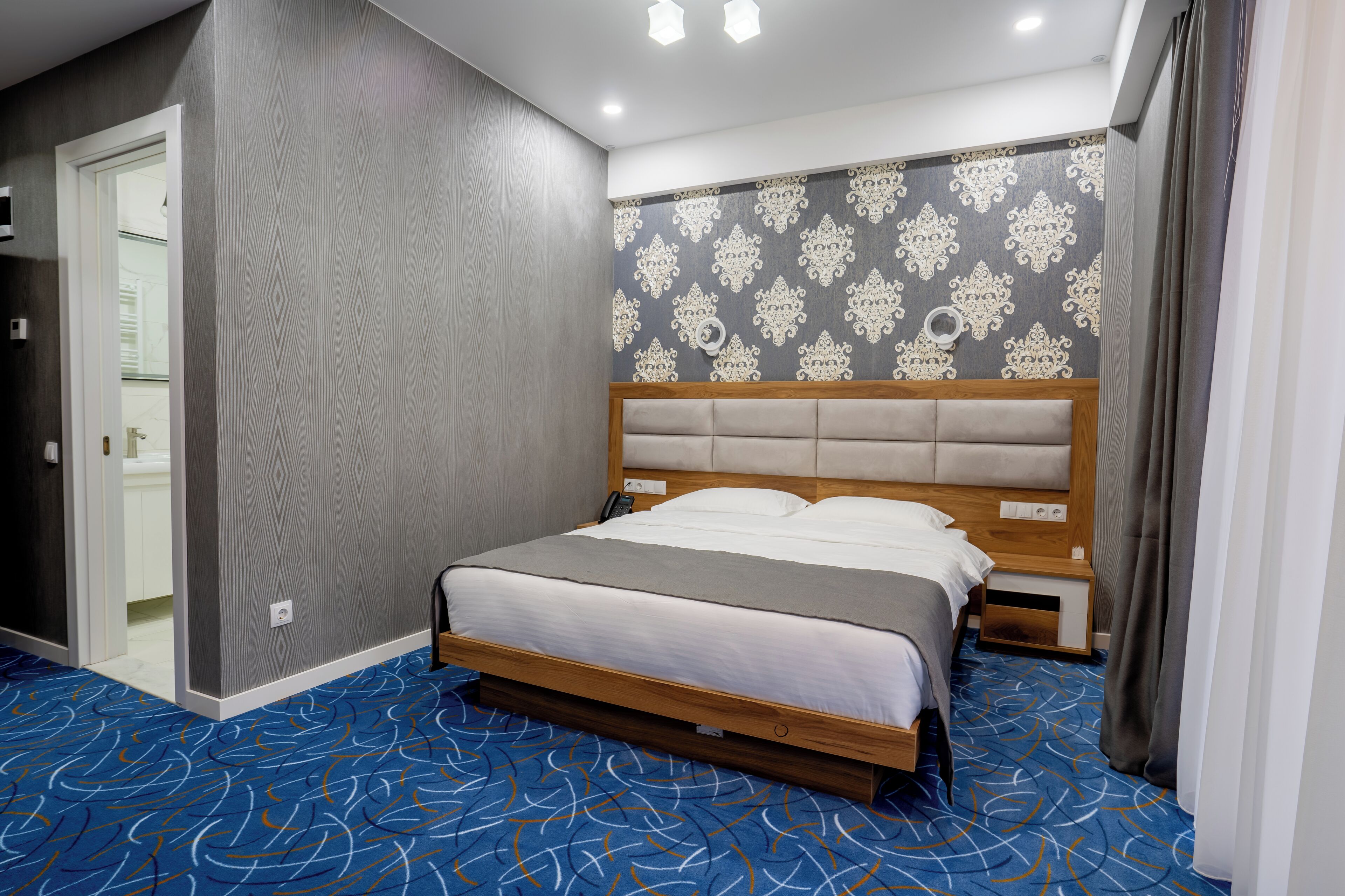 Quarto de hotel moderno com uma cama grande, cabeceira de madeira, parede estampada e tapete azul.