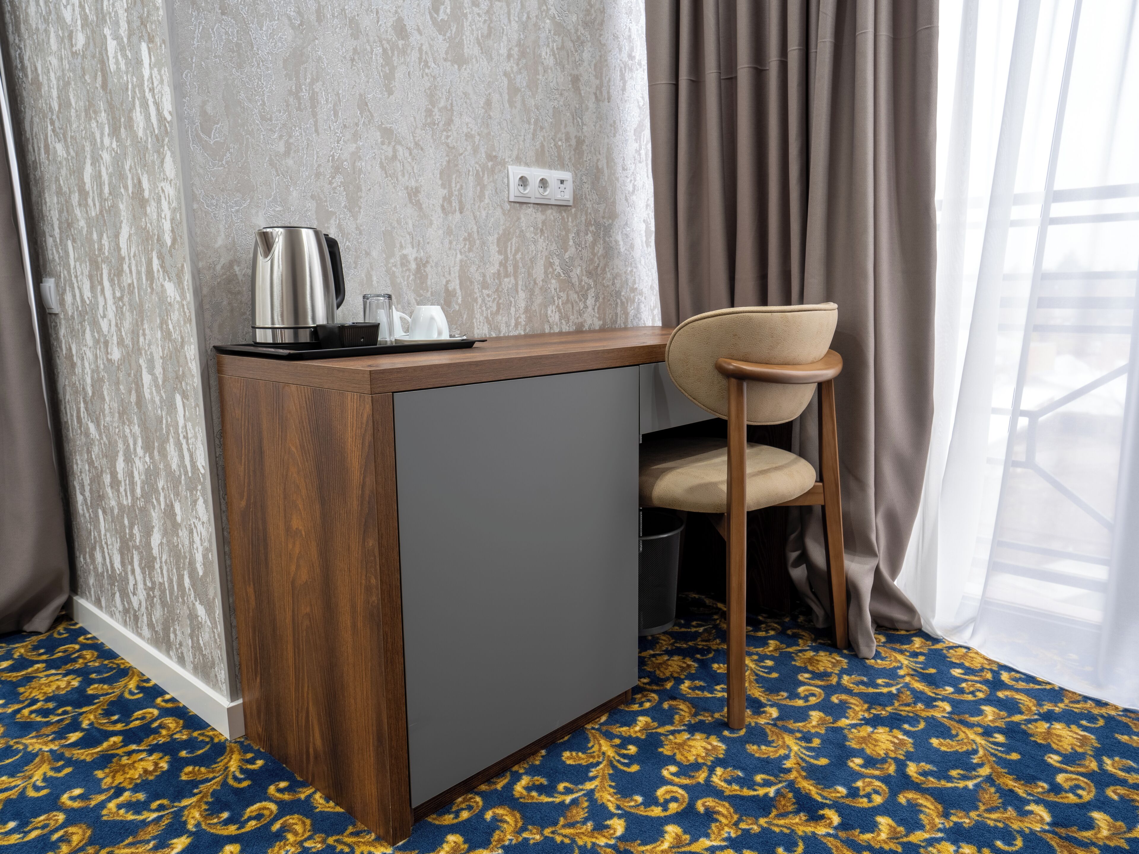 A compact room with a wooden desk, a grey cabinet, a beige chair, and a patterned carpet.