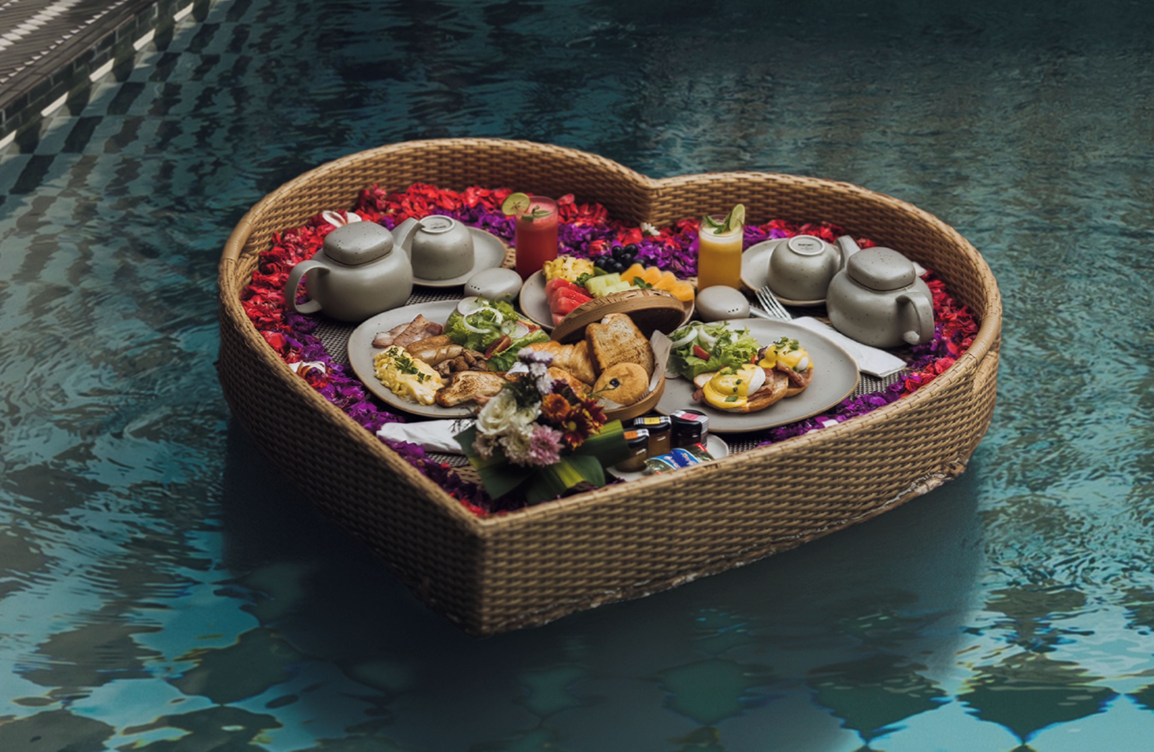 A heart-shaped basket filled with food and drinks on a reflective surface.