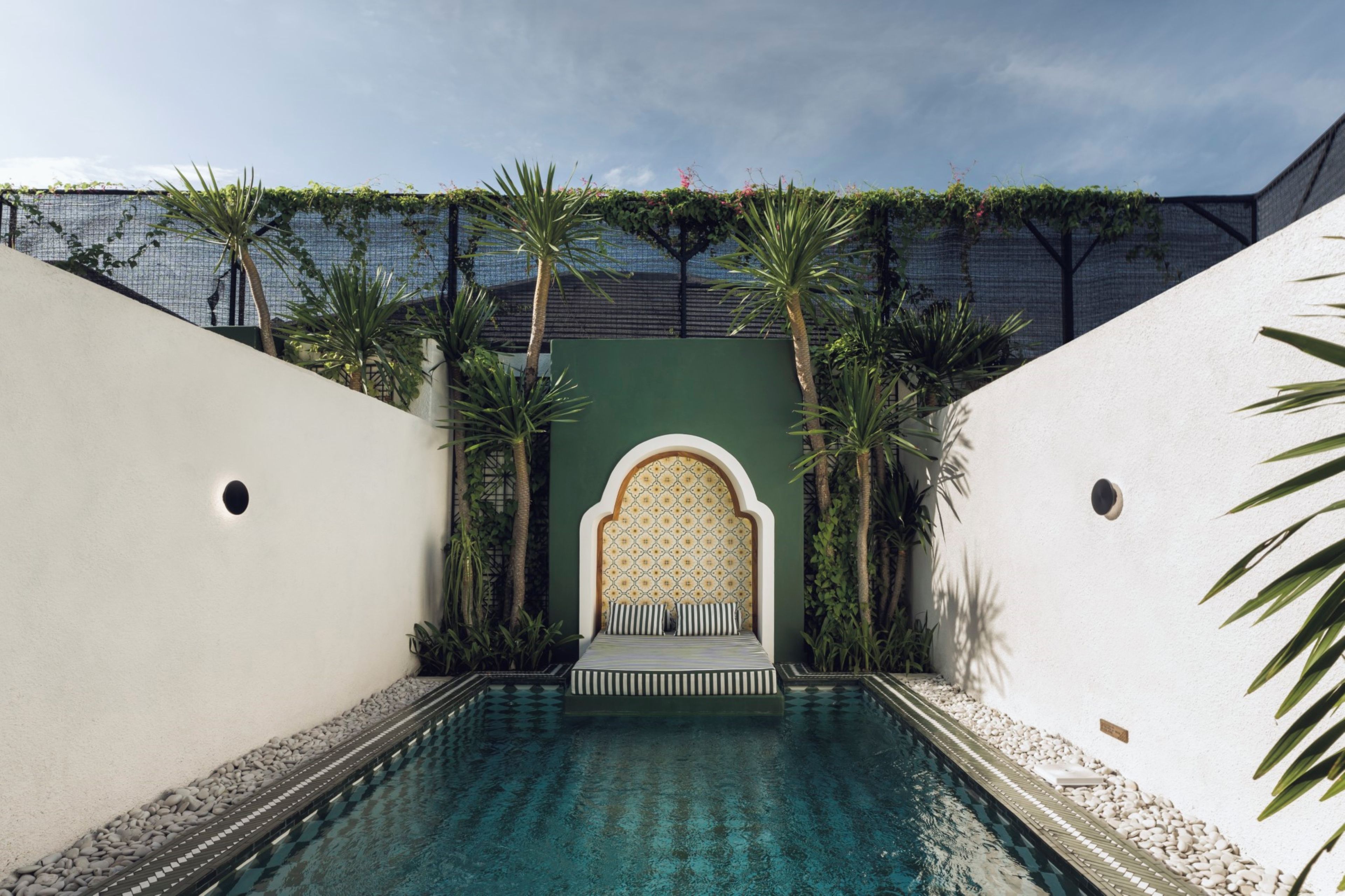 A pool area with a seating area, surrounded by greenery and a patterned wall.
