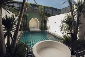 A swimming pool with a sunken seating area, surrounded by tropical plants and a white wall with a decorative arch. - Palmea Villa Seminyak by Ini Vie Hospitality (Seminyak)