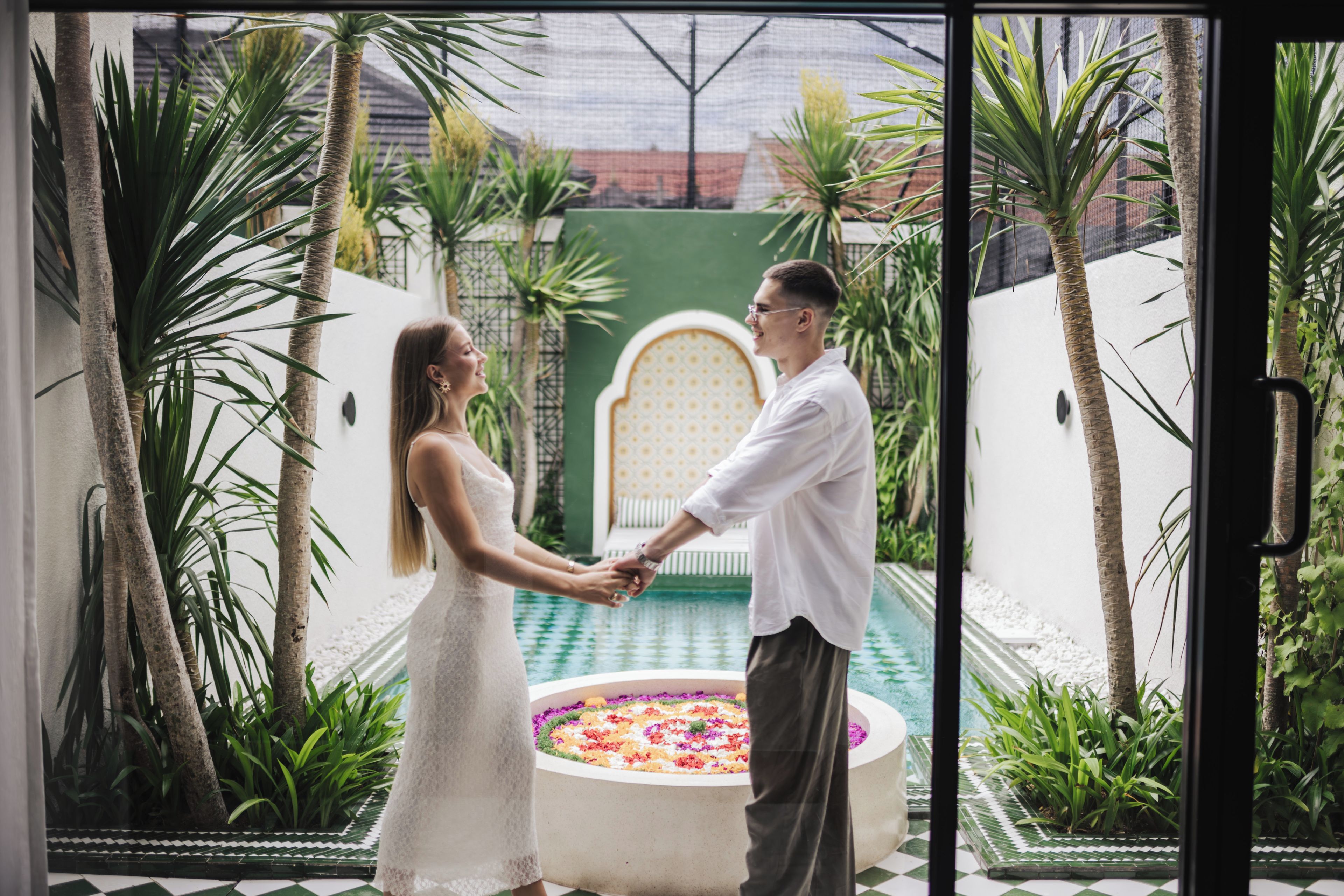 Un couple se tenant la main dans une cour ornée d’une piscine décorative et de plantes tropicales.