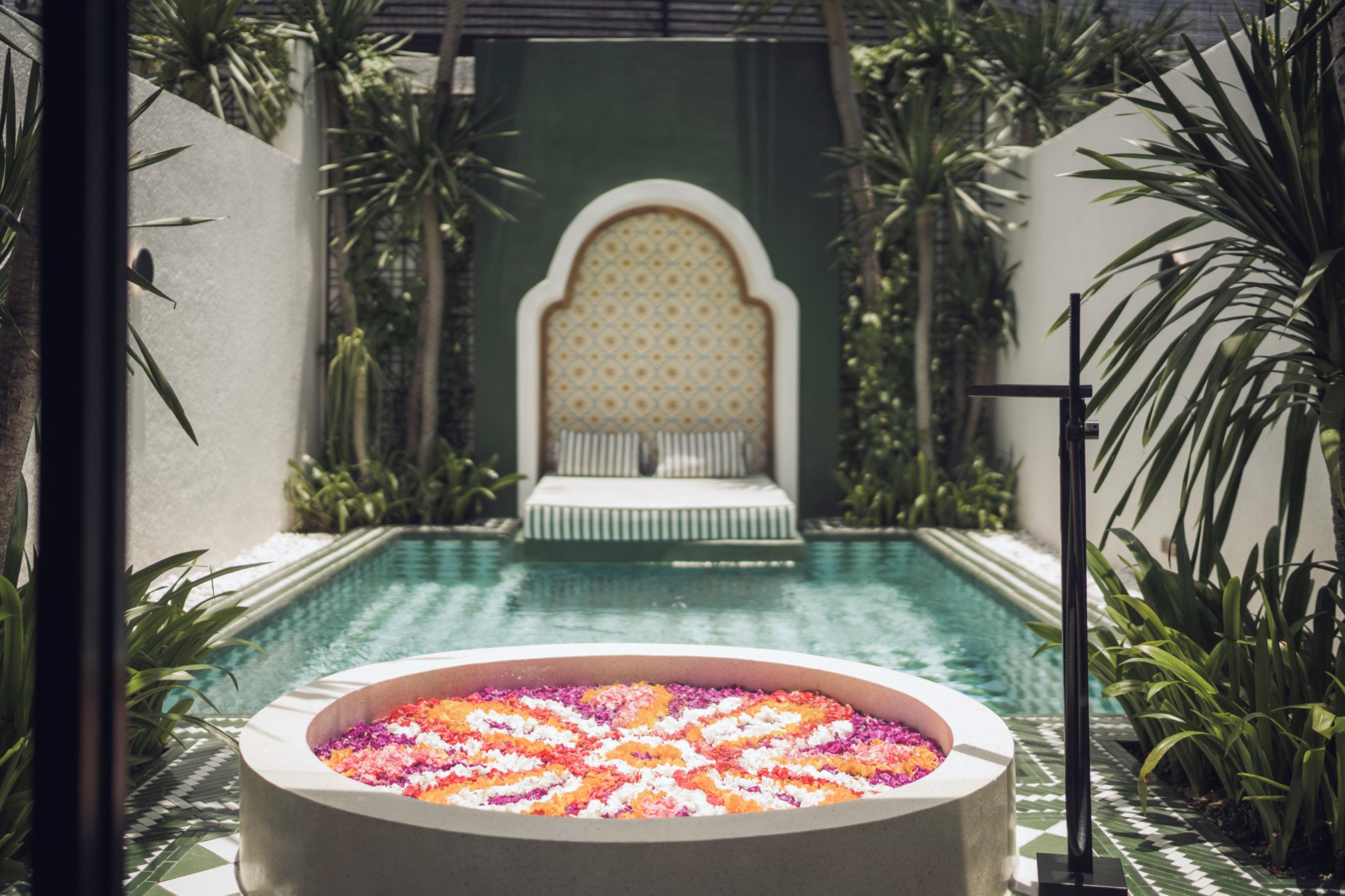 A pool area with a circular flower arrangement in the foreground, a bench with a patterned backrest in the background, and tropical plants surrounding the space.