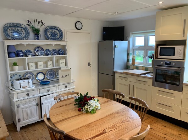 Une cuisine avec des meubles blancs, une table à manger en bois avec des chaises, un réfrigérateur, un four à micro-ondes et une horloge au mur.