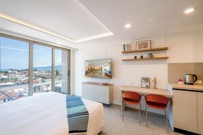 A hotel room with a bed, a desk, two chairs, a mirror, and a view of the city. - HOMA Chalong Bay (Chalong)