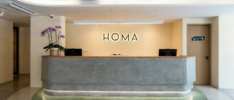 A reception desk with 'HOMA' signage, a potted orchid, and a green carpet.