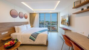 A modern hotel room with a large bed, a desk, a chair, a TV, and a view of the city. - HOMA Chalong Bay (Chalong)
