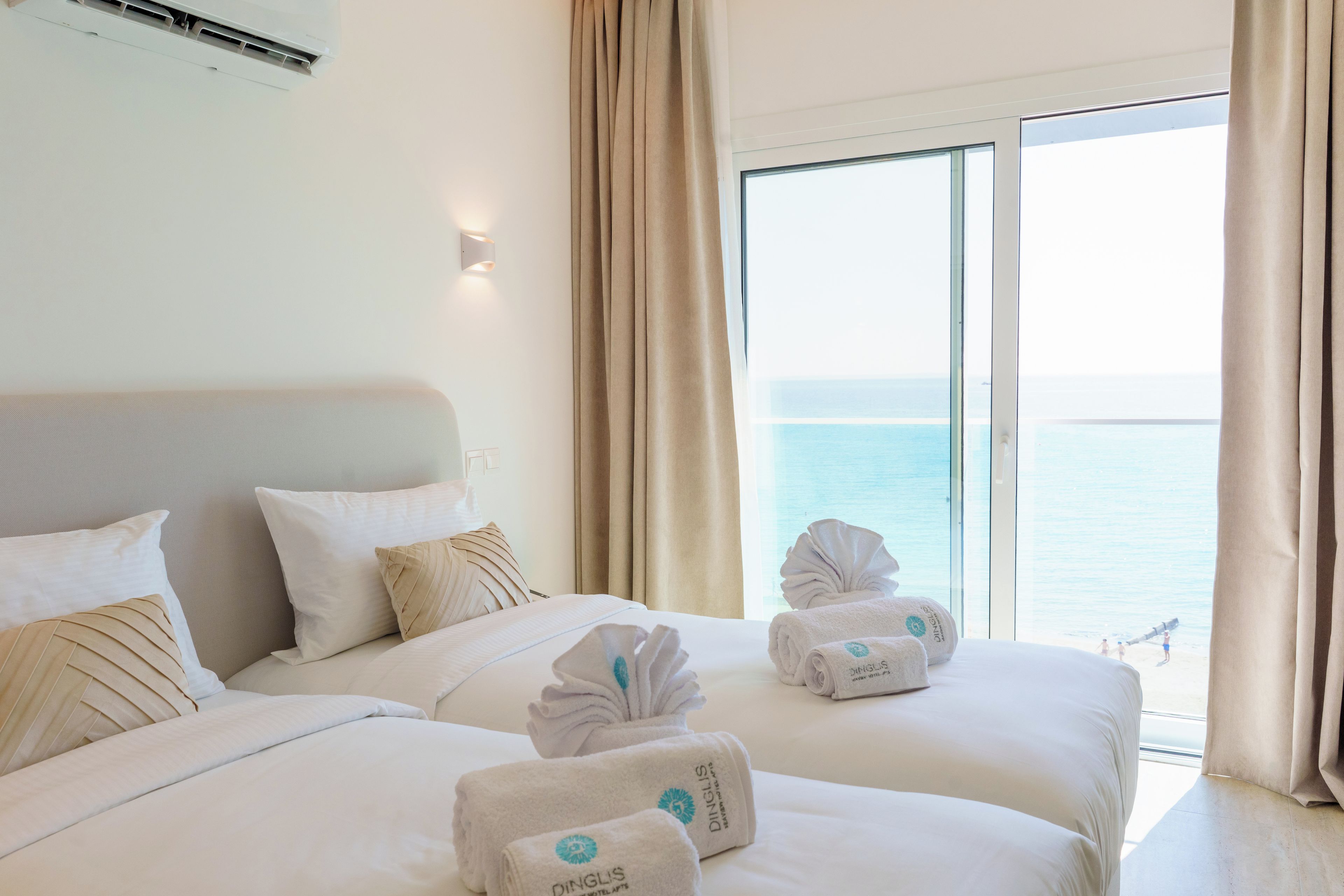 A hotel room with a bed, pillows, a wall-mounted air conditioner, and a view of the sea through a large window.