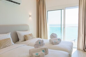 A hotel room with a bed, pillows, a wall-mounted air conditioner, and a view of the sea through a large window. - Dinglis Beach Hotel Suites (Larnaca)