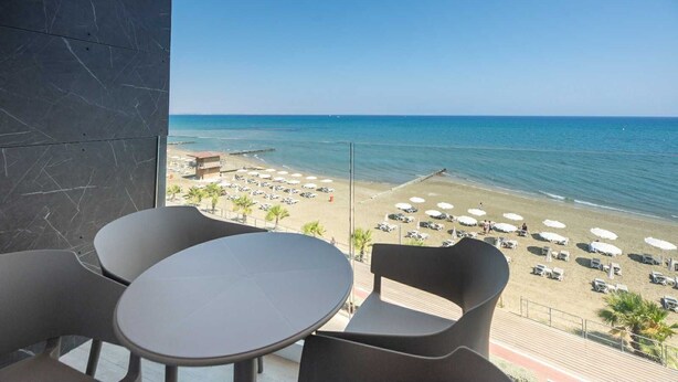 A balcony with a view of a beach, featuring a table and chairs, and a building with a pool. - Dinglis Beach Hotel Apartments (Larnaca)