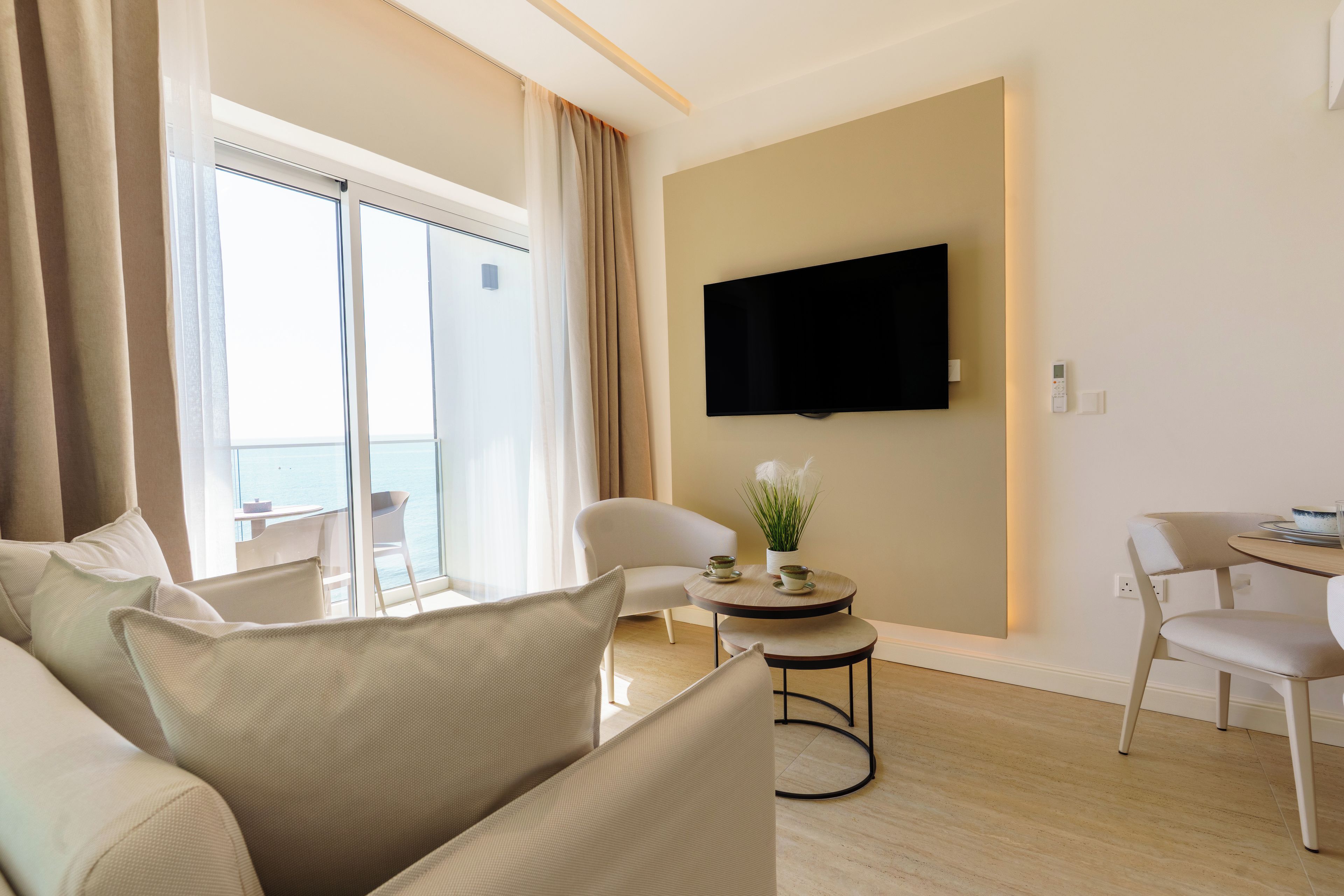 Une chambre d’hôtel moderne dotée d’un balcon, d’un téléviseur à écran plat fixé au mur, d’une table de salle à manger ronde avec des chaises et d’un canapé avec des coussins.
