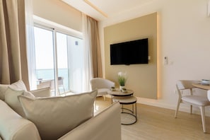 A modern hotel room with a balcony, a flat-screen TV mounted on the wall, a round dining table with chairs, and a sofa with cushions. - Dinglis Beach Hotel Suites (Larnaca)