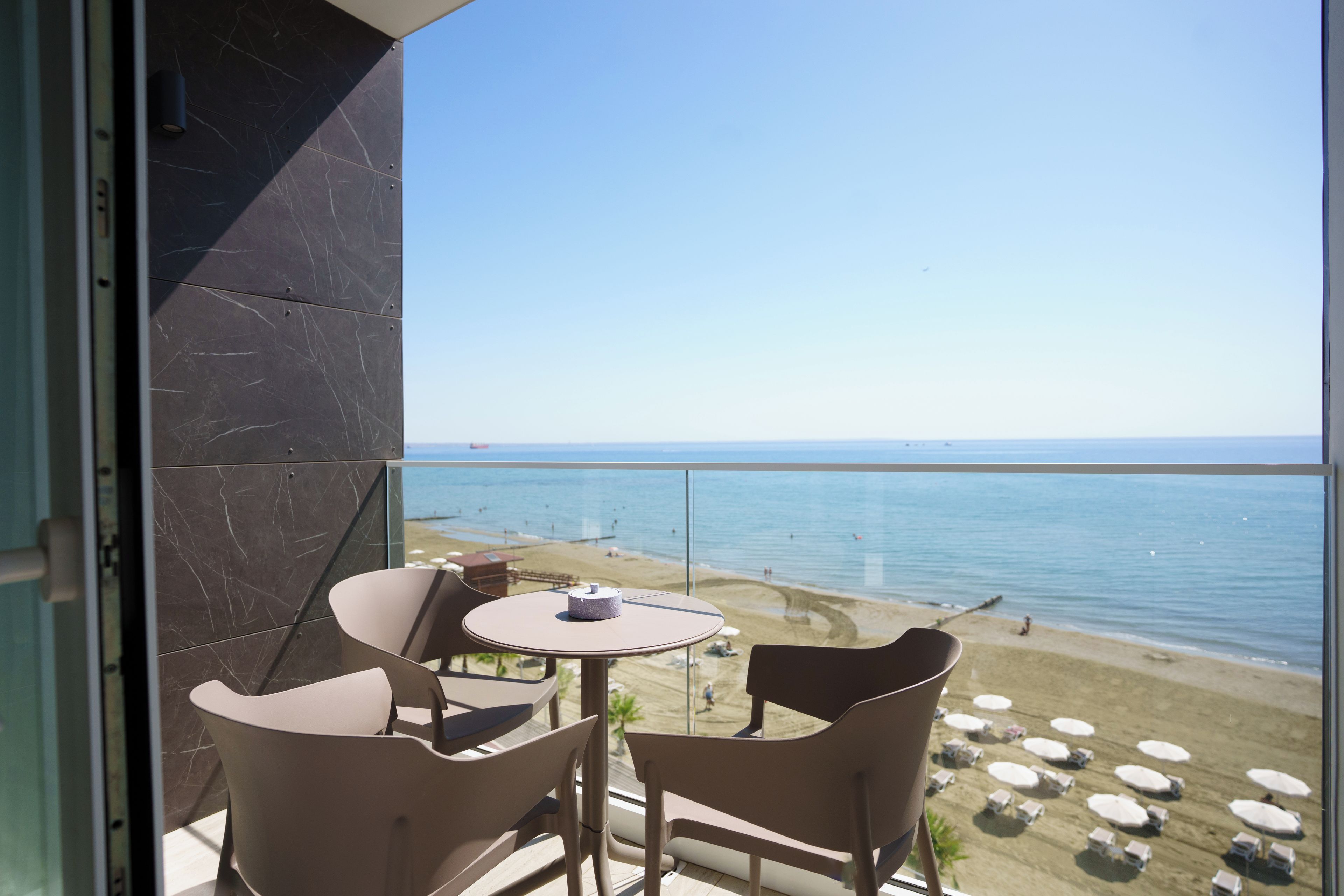 Varanda com mesa e cadeiras com vista para uma praia com guarda-sóis e pessoas.