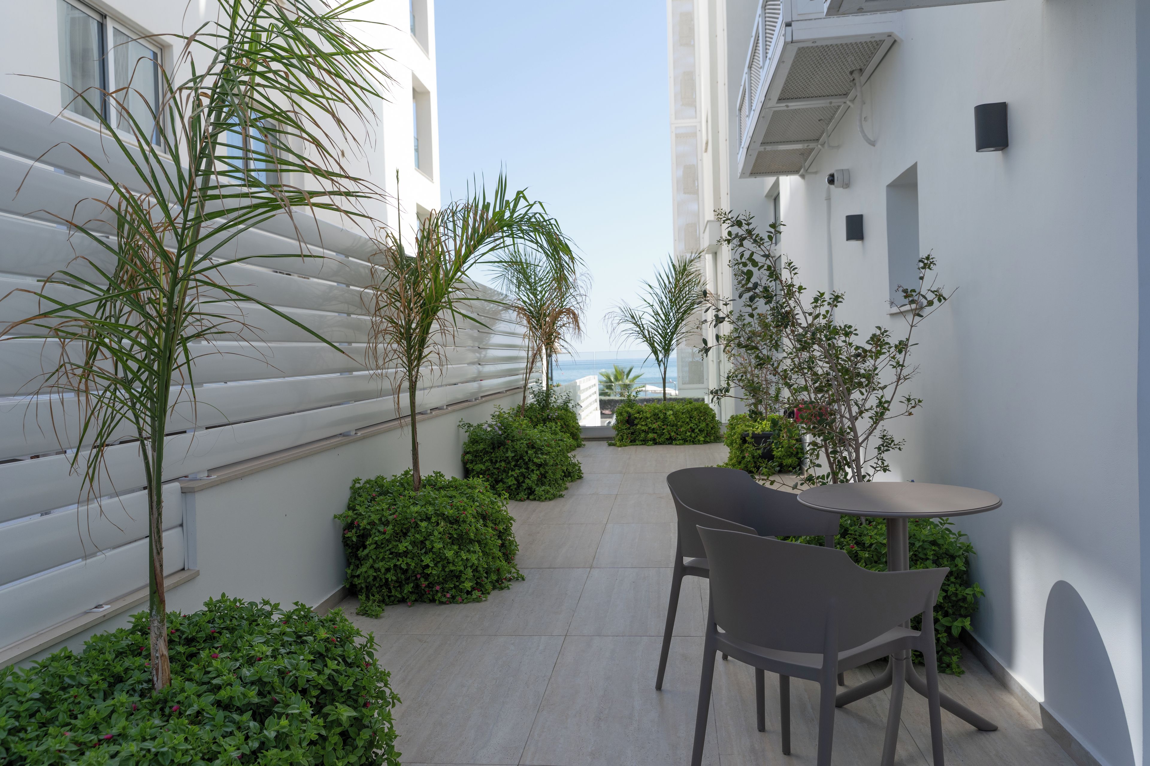 A modern outdoor patio area with a table and chairs, surrounded by greenery and a view of the sea.