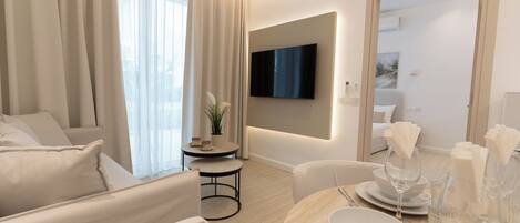 A modern living room with a flat-screen TV mounted on the wall, a dining area with a table set for two, and a seating area with a sofa and armchairs.
