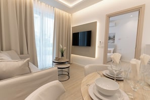 A modern living room with a flat-screen TV mounted on the wall, a dining area with a table set for two, and a seating area with a sofa and armchairs. - Dinglis Beach Hotel Suites (Larnaca)