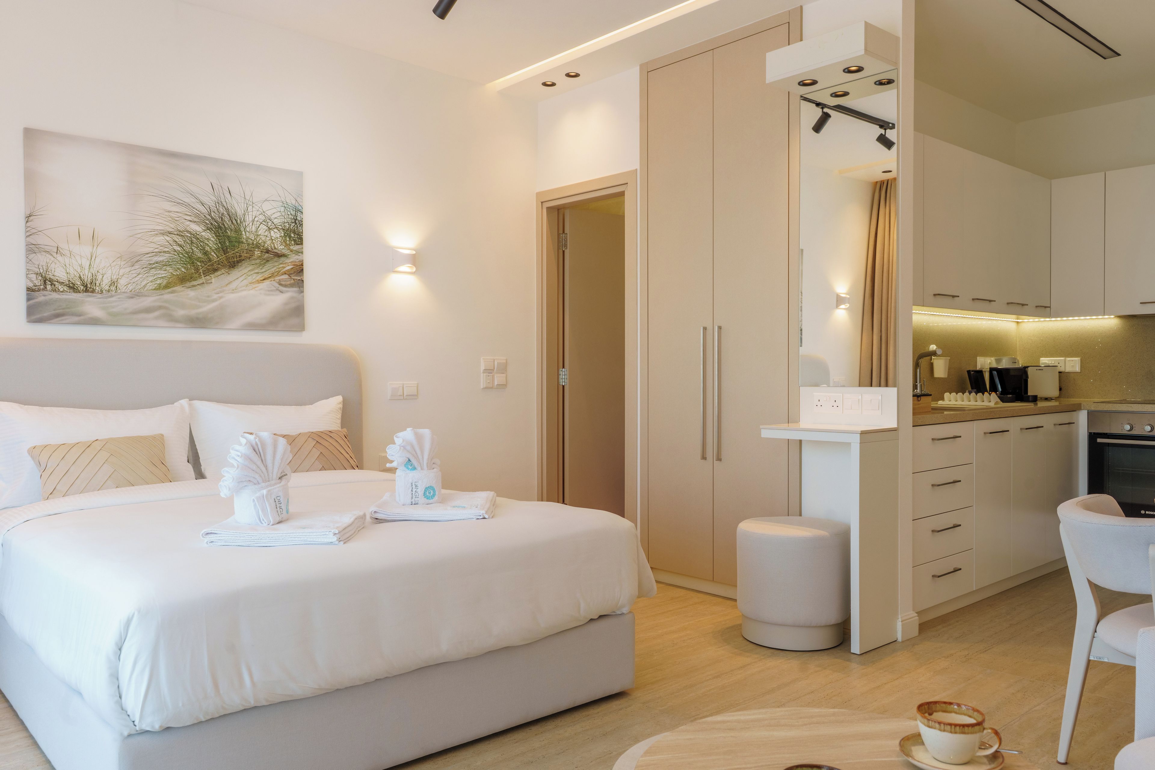 A modern hotel room with a bed, a small seating area, a kitchenette, and a bathroom.