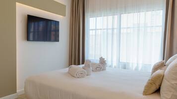 Habitación de hotel con una cama grande, un televisor de pantalla plana montado en la pared y un ventanal con cortinas translúcidas.