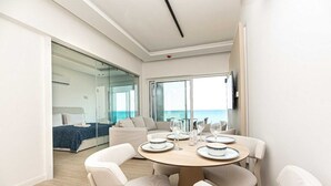 A modern room with a round dining table, white chairs, a sofa, and a large window with a view of the sea. - Dinglis Beach Hotel Apartments (Larnaca)