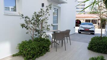 Un área de estar moderna al aire libre con una mesa y sillas, rodeada de follaje y una fachada de edificio blanca.