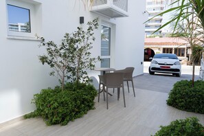 A modern outdoor seating area with a table and chairs, surrounded by greenery and a white building facade. - Dinglis Beach Hotel Suites (Larnaca)