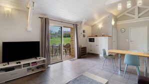A modern living room with a flat-screen TV, a dining table with chairs, a kitchen area, and a sliding glass door leading to a garden. - Holiday Home “La Cense De La Mérelie - Le Logis” with Shared Pool, Wi-Fi and Air Conditioning (Saint-Geniès)