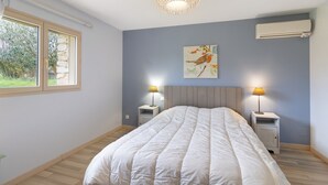 A bedroom with a bed, bedside lamps, a wall-mounted air conditioner, and a window with a view of greenery. - Holiday Home “La Cense De La Mérelie - Le Logis” with Shared Pool, Wi-Fi and Air Conditioning (Saint-Geniès)